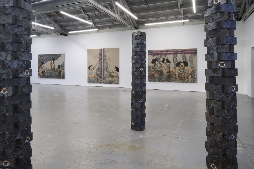 a gallery with paintings on the wall and three large freestanding sculptural poles made of bricks painted black