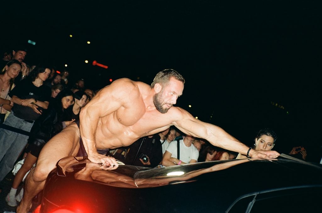 a body builder climbs atop a car as an audience looks on in a performance art piece by Göksu Kunak 