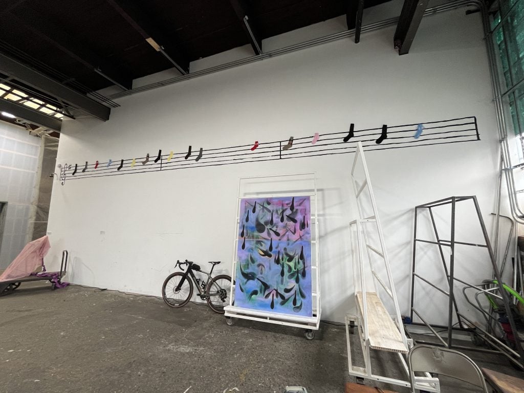 In a color photo, a musical scale is high on a white wall. A bike leans against it, near an abstract painting of many colors.