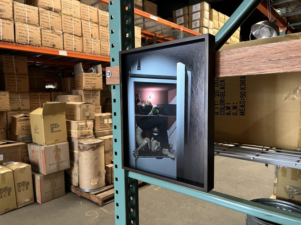 In a color photo, a photo in a black frame is hanging on the edge of a shelf in a large warehouse. The photo shows a doll-like figure stuck inside a refrigerator.