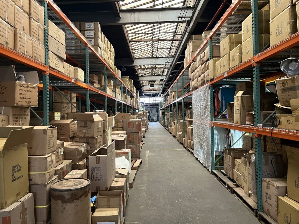 A large warehouse with tall ceilings and skylights is seen in a color photo. Four tall shelves are mostly filled with cardboard boxes.