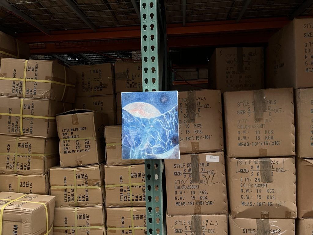 In a color photo, cardboard boxes are stacked in a warehouse and a small painting—mostly blue and vaguely abstract—hangs on a thin metal column.