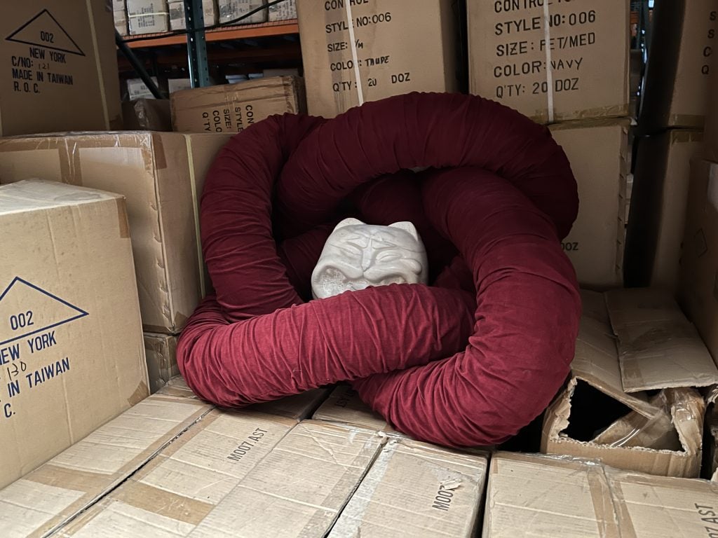A white sculpture of a small, smiling cat is nestled in a burgundy pillow-like form in this color photo. It sits amid cardboard boxes.