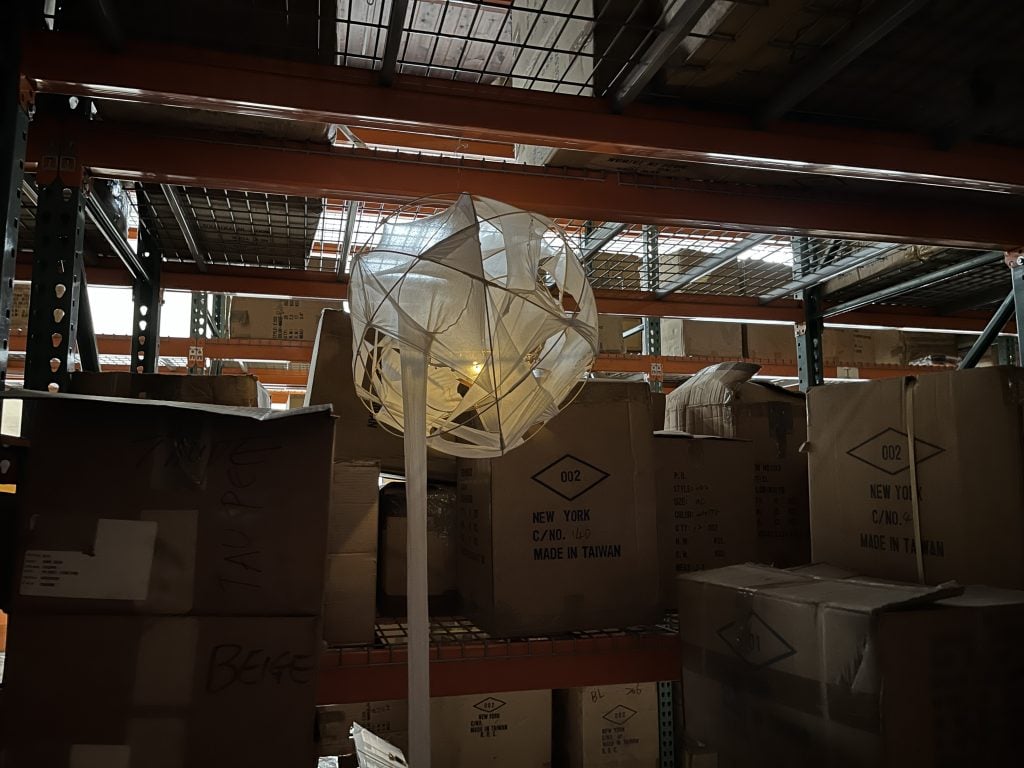 In a dimly lit space, amid cardboard boxes, hangs a spherical sculpture covered with white fabric. Inside, a digital candle glows.