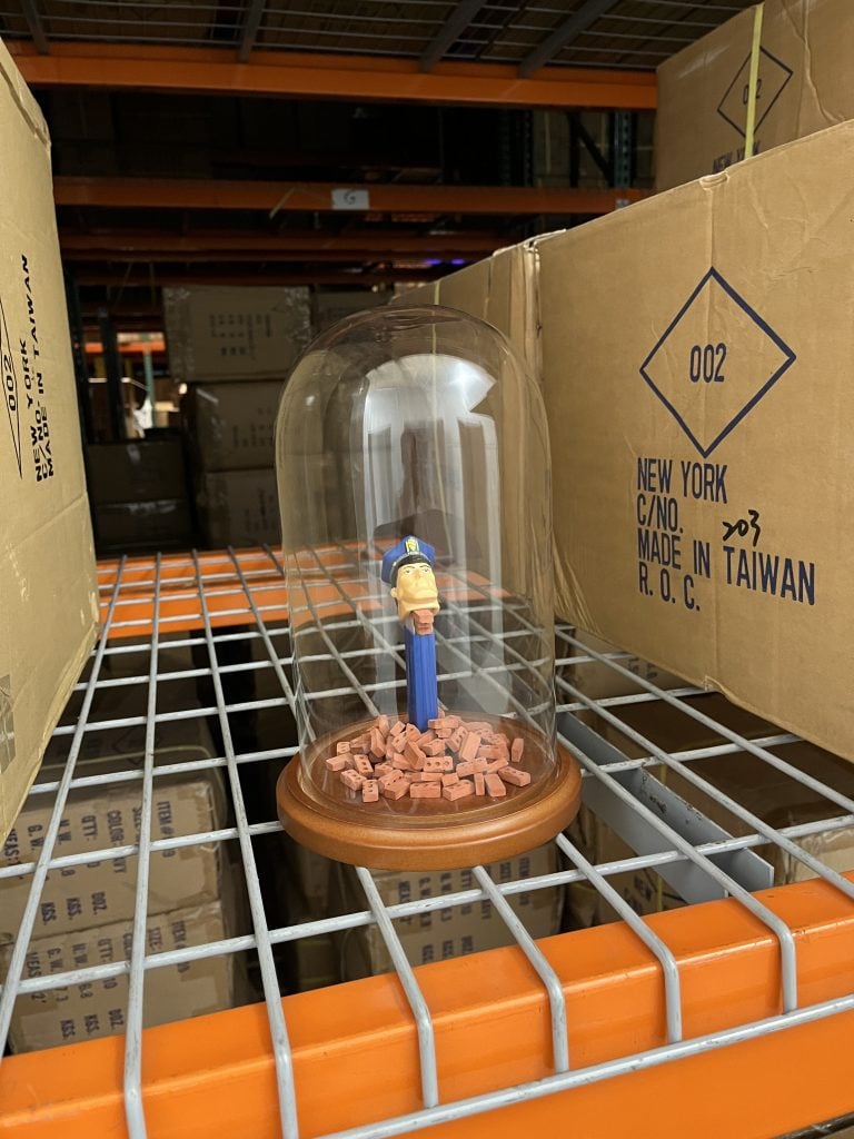 In a color photo, a Pez candy dispenser sits under a glass dome and spits out tiny bricks. It is atop a wire rack surrounded by cardboard boxes.