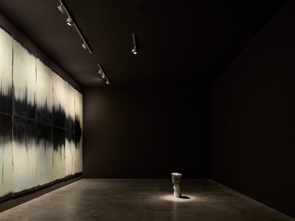 a very dark big exhibition space with a large long monochromatic painting with a stretch of black inside a large white rectangle runs along one wall. An urn like monument sits on the floor under a spotlight.