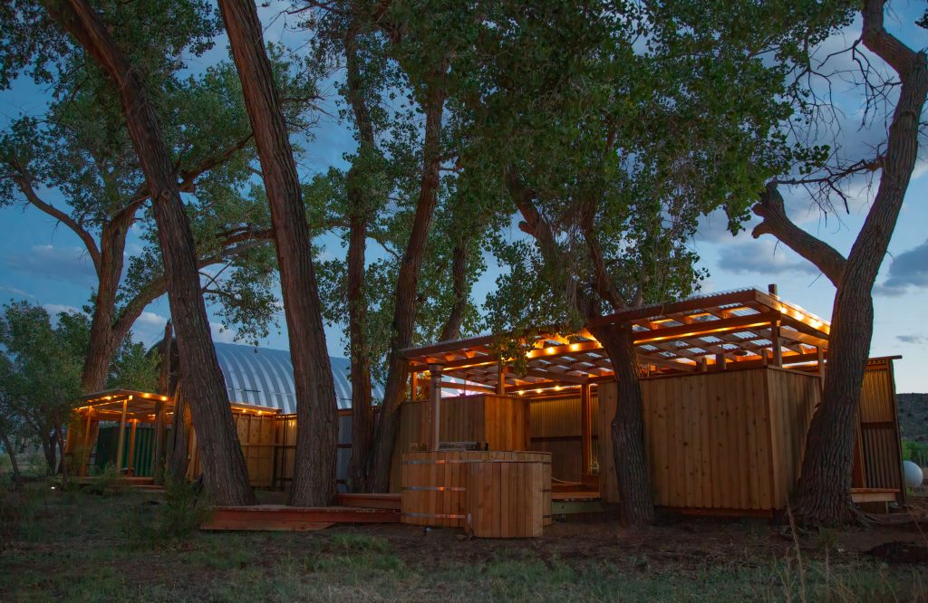 A cozy outdoor space with wooden structures and a hot tub, illuminated by warm lights, nestled among tall trees at dusk.