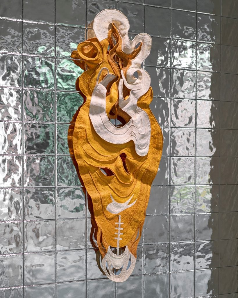 A loopy orange and cream textile work hangs on a silver wall.