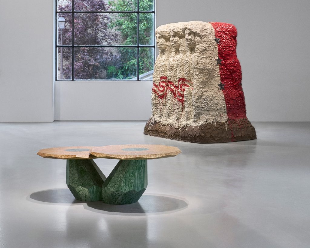 A ceramic sculpture of three women's faces carved into a mound with red and white dripping glaze, in the foreground a set of two round tables shaped like mushrooms