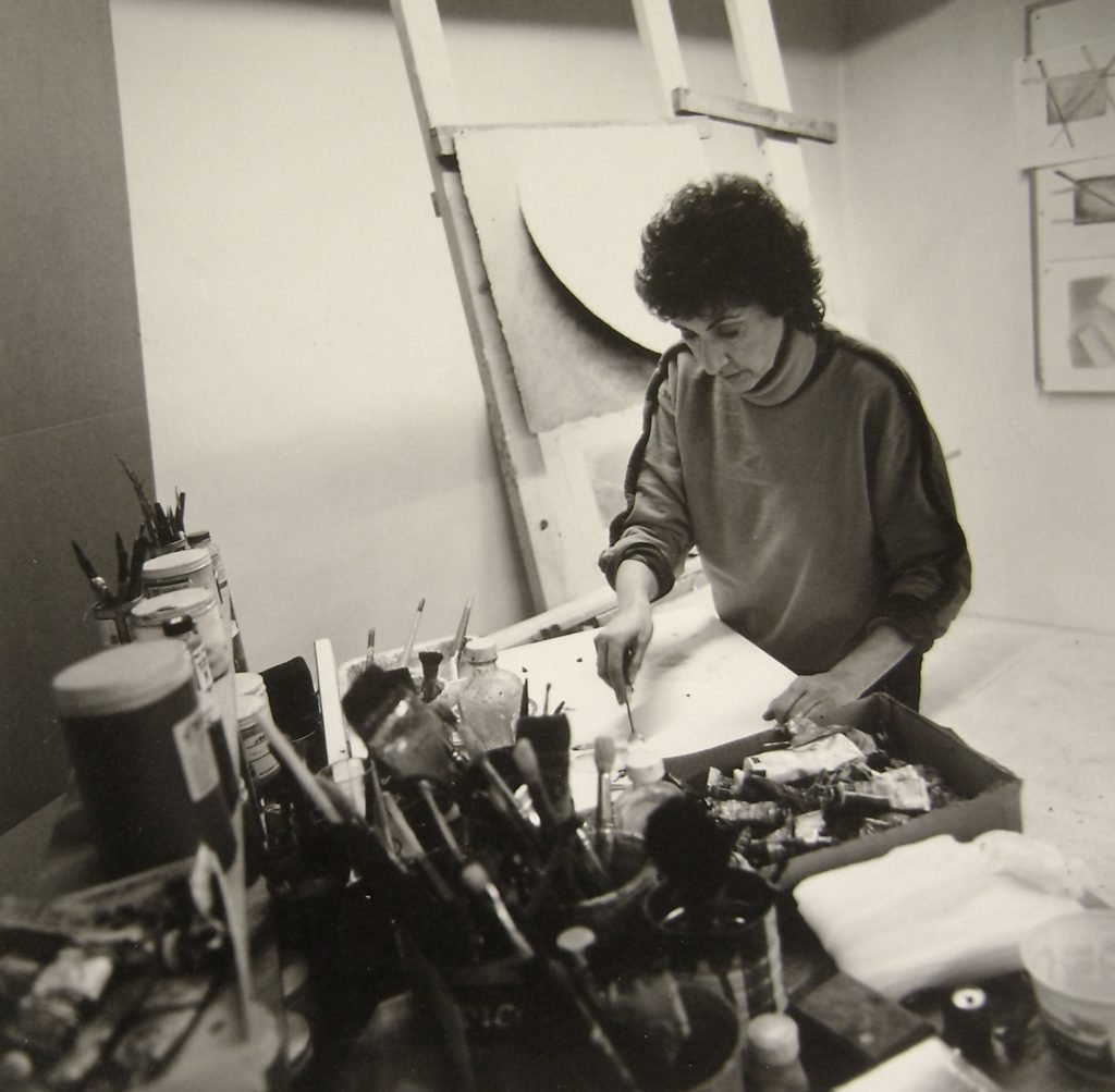 a black and white photo in which a woman stands in an artist's studio with a brush in her hands surrounded by brushes and paint