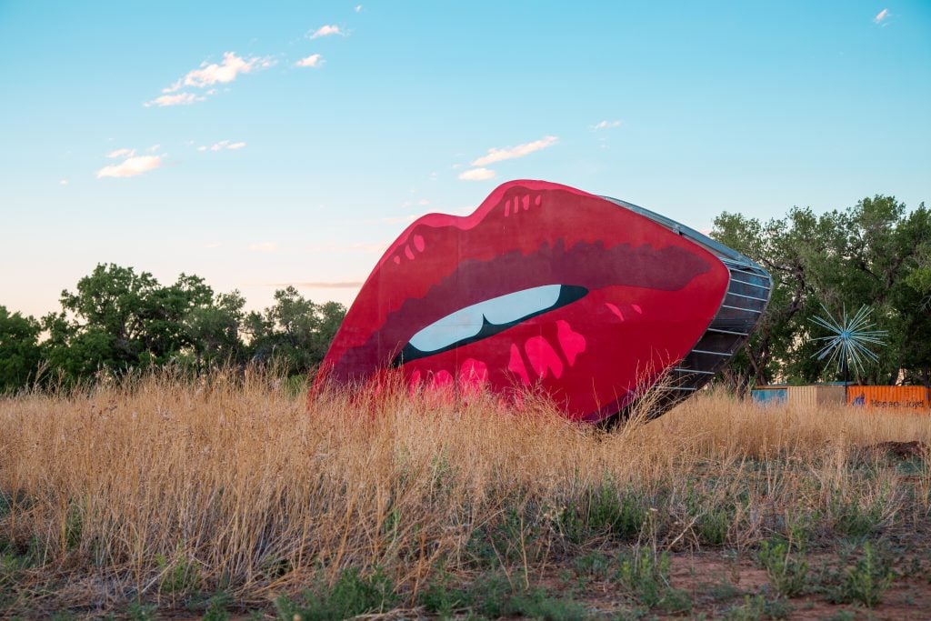 A large, outdoor art installation featuring bright red lips in a grassy field with trees and a clear blue sky in the background.
