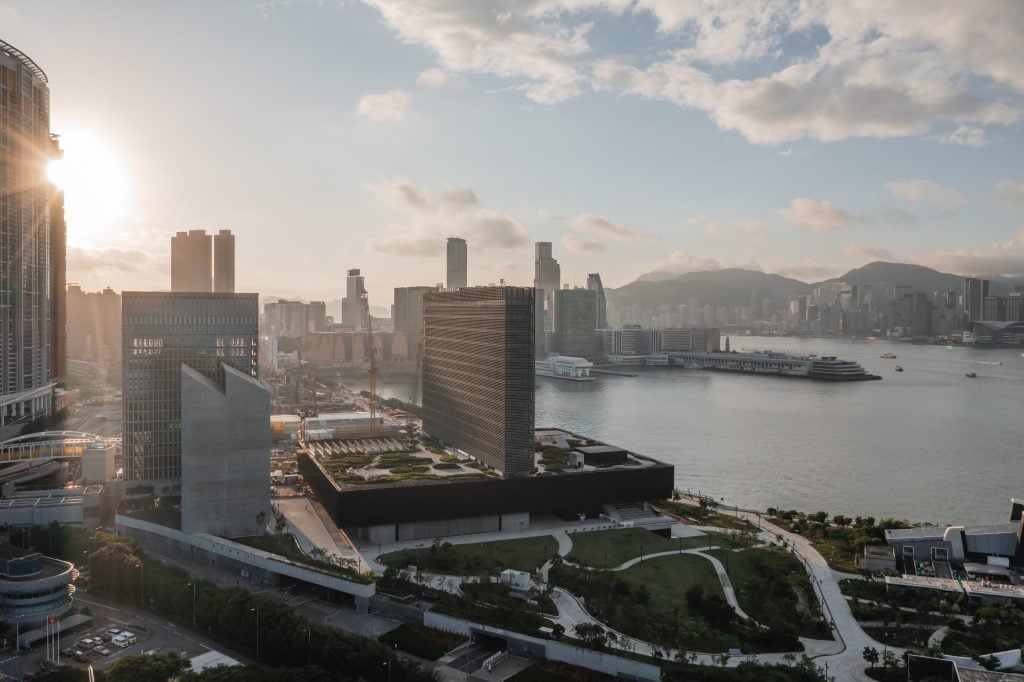 an image of M+ museum in Hong Kong taken from the sky. tall buildings can be seen in the foreground, hills in the background. it is sunny with clouds