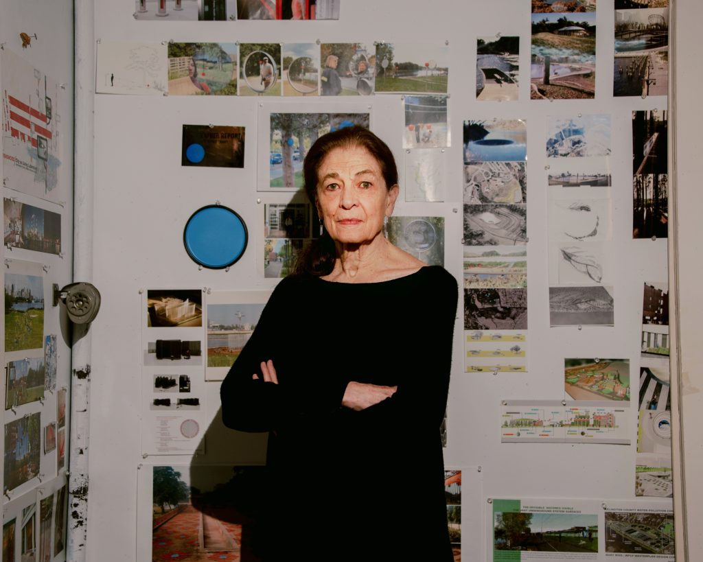 A woman stands confidently in her studio surrounded by various project images and notes pinned to a wall, indicating a creative or planning process. She is dressed in a simple black outfit and has a contemplative expression.