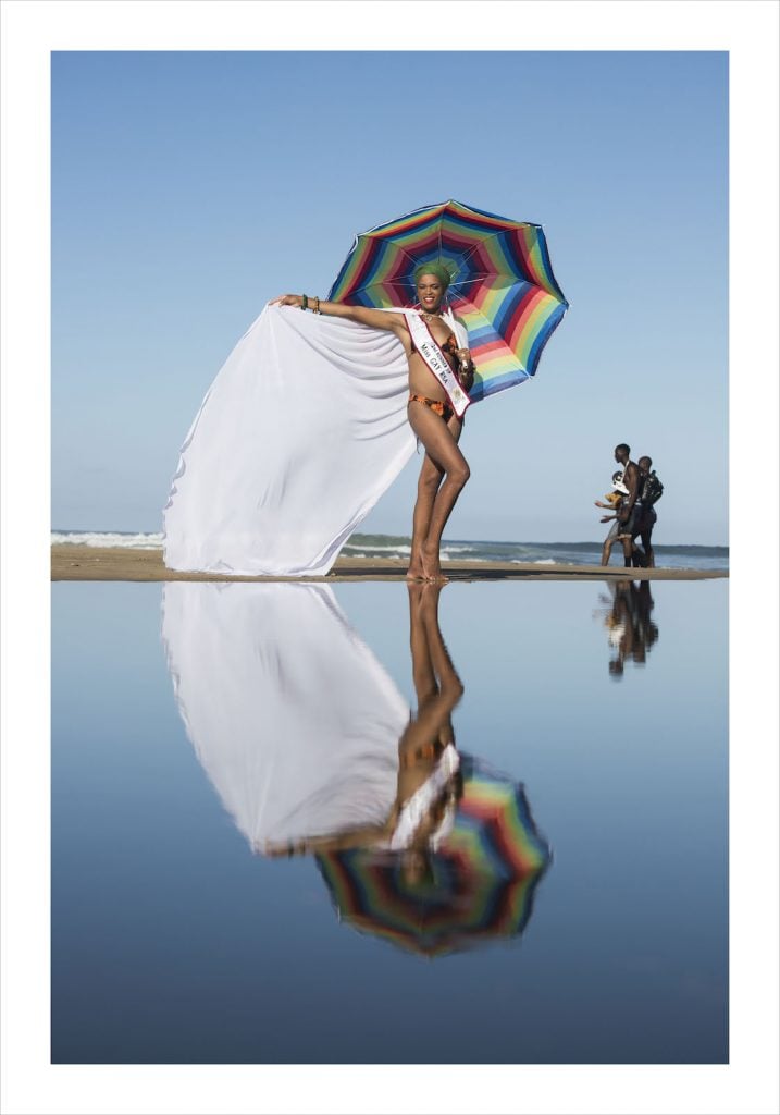 a person wearing a sash and a white cape holds a rainbow umbrella. their reflection is seen in the water. By artist Zanele Muholi.