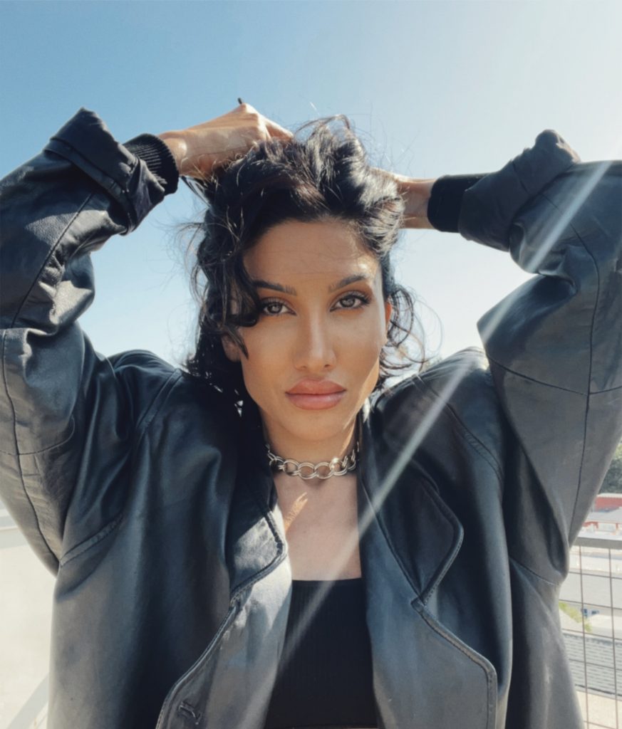 A portrait of a woman outdoors, her hands lifting her curly hair, wearing a black leather jacket and a chunky chain necklace. The sunlight highlights her face against a clear blue sky.