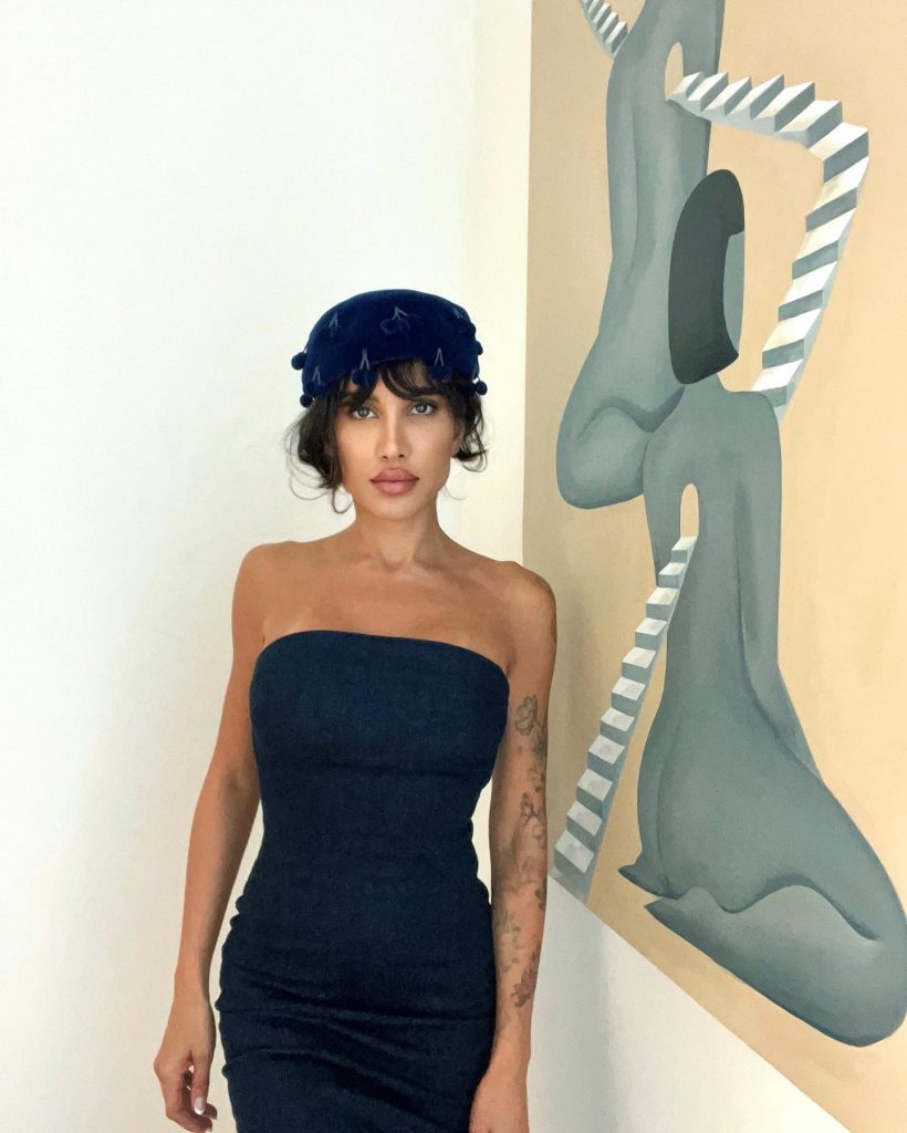A woman in a dark strapless dress and a navy blue beret, with subtle tattoos visible on her arms, standing next to a painting of a stylized nude figure entwined with black and white striped elements.
