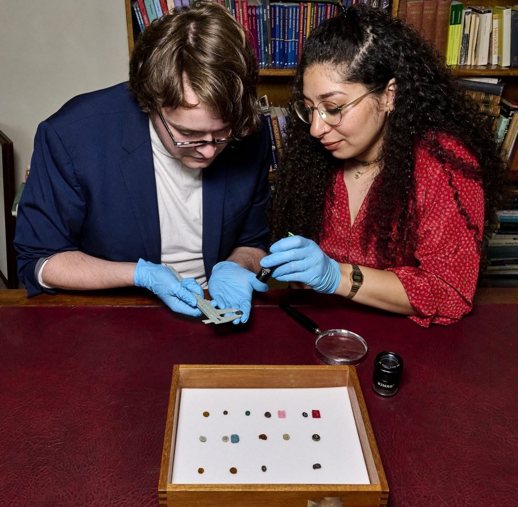 a man and a woman look closely at a gem together while wearing blue globes, a tray of several more gems sits on the table in front of them