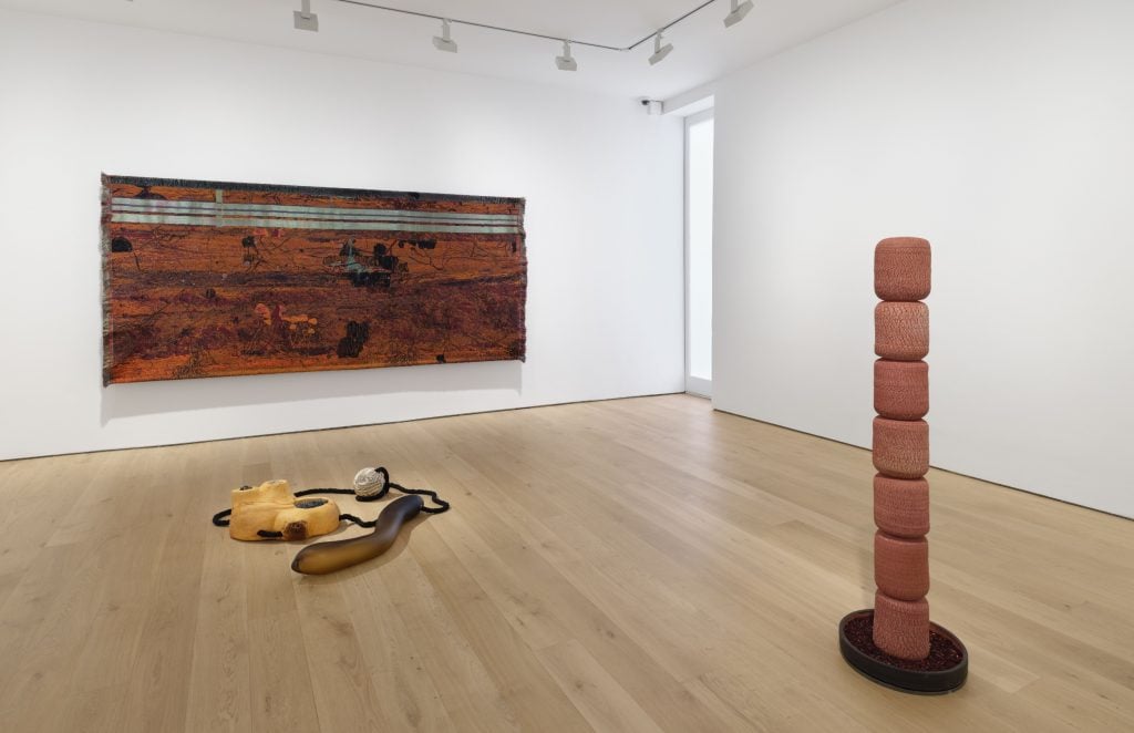 a brown textile hangs on a gallery wall behind two sculptures, one is a floor sculpture with shapes connected by rope and the other is a tall sculpture reaching up from the floor. 