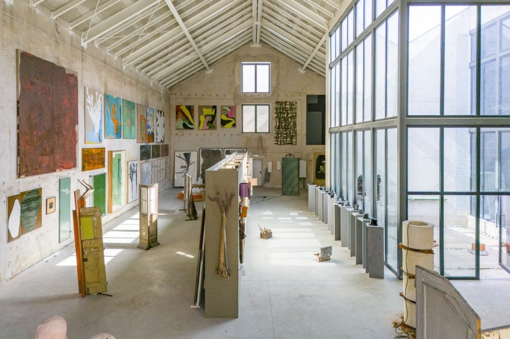 a large spacious warehouse type room with lots of paintings hanging on the wall at all heights. some sculptural works fill the inside space.
