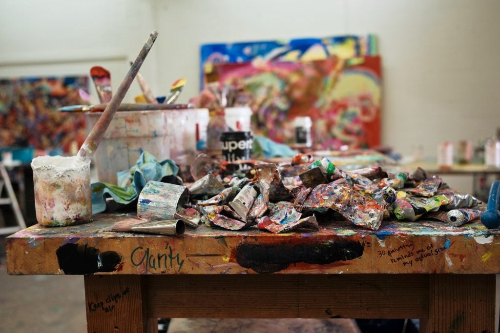 A messy table is covered in paint brushes, tubes and buckets. In a studio belonging to Lauren Quin