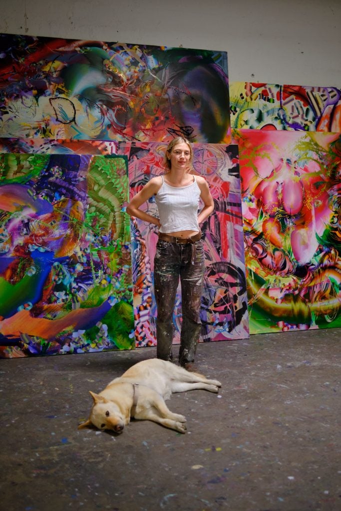 A woman in a white vest and grey trousers stands infant of several colourful abstract paintings. A blond dog lies at her feet.