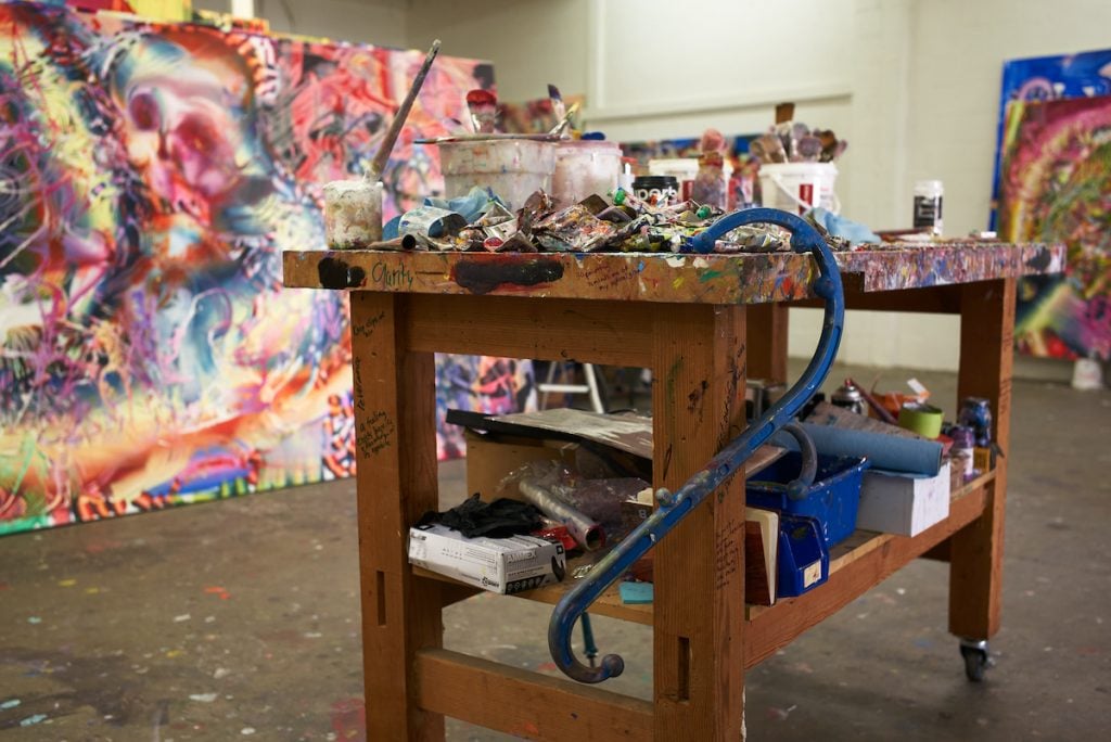 A messy table is covered in paint brushes, tubes and buckets. In a studio belonging to Lauren Quin
