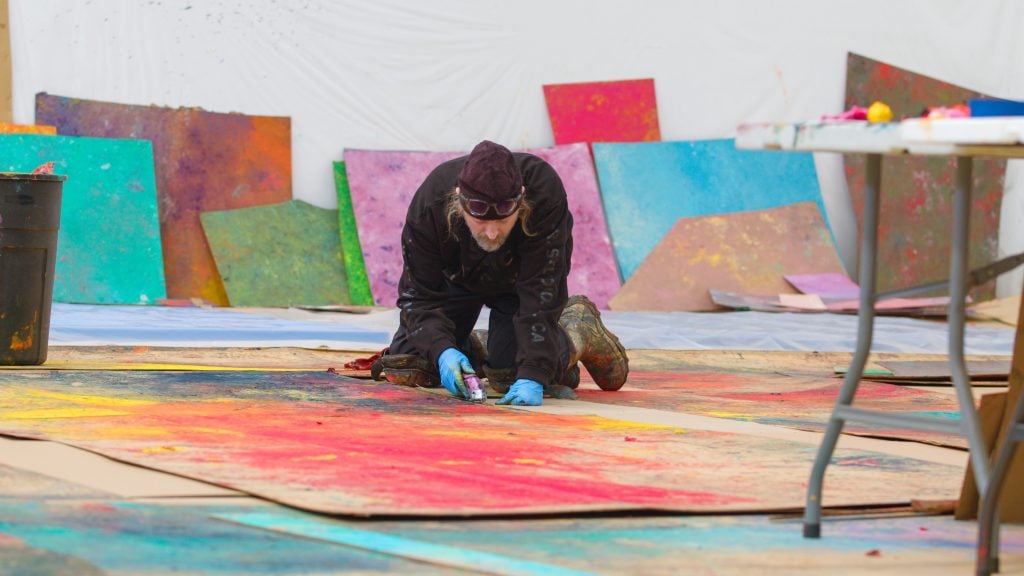 an artist crouches on the floor working on paintings