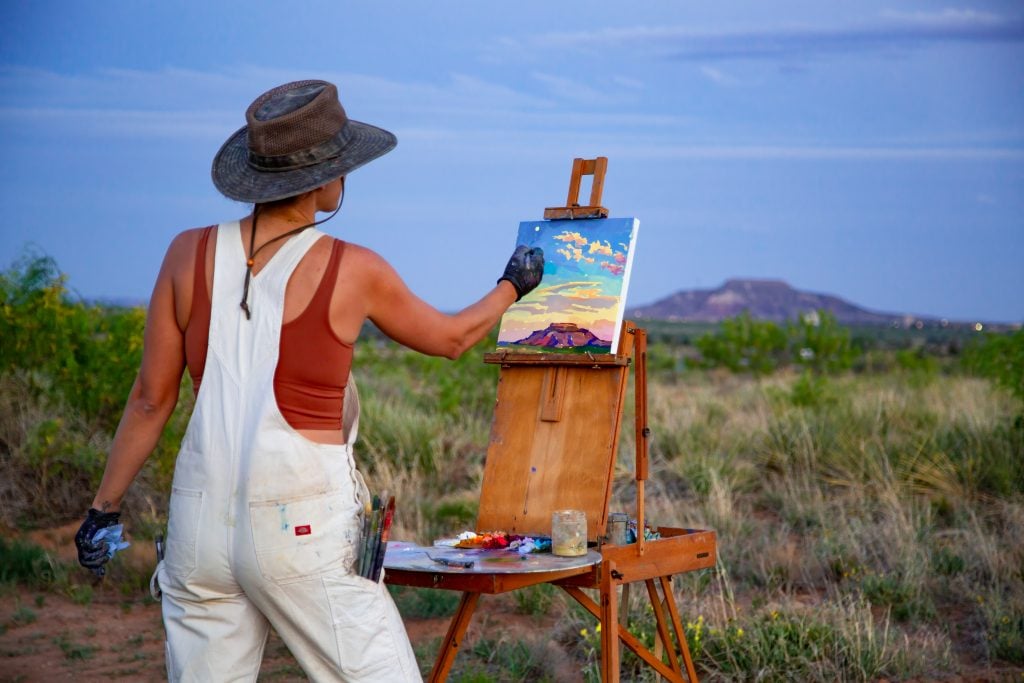 An artist wearing a hat and overalls paints a colorful landscape on an easel in a grassy field during golden hour, with mountains visible in the background.