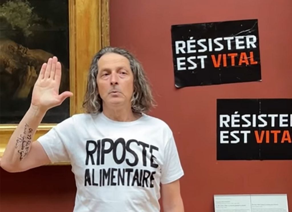 A man stands in front of a painting in a museum, raising his right hand in a gesture. He is wearing a white T-shirt with the words "Riposte Alimentaire" and the phrase "Résister est vital" repeated on his shirt and on black signs on the wall.