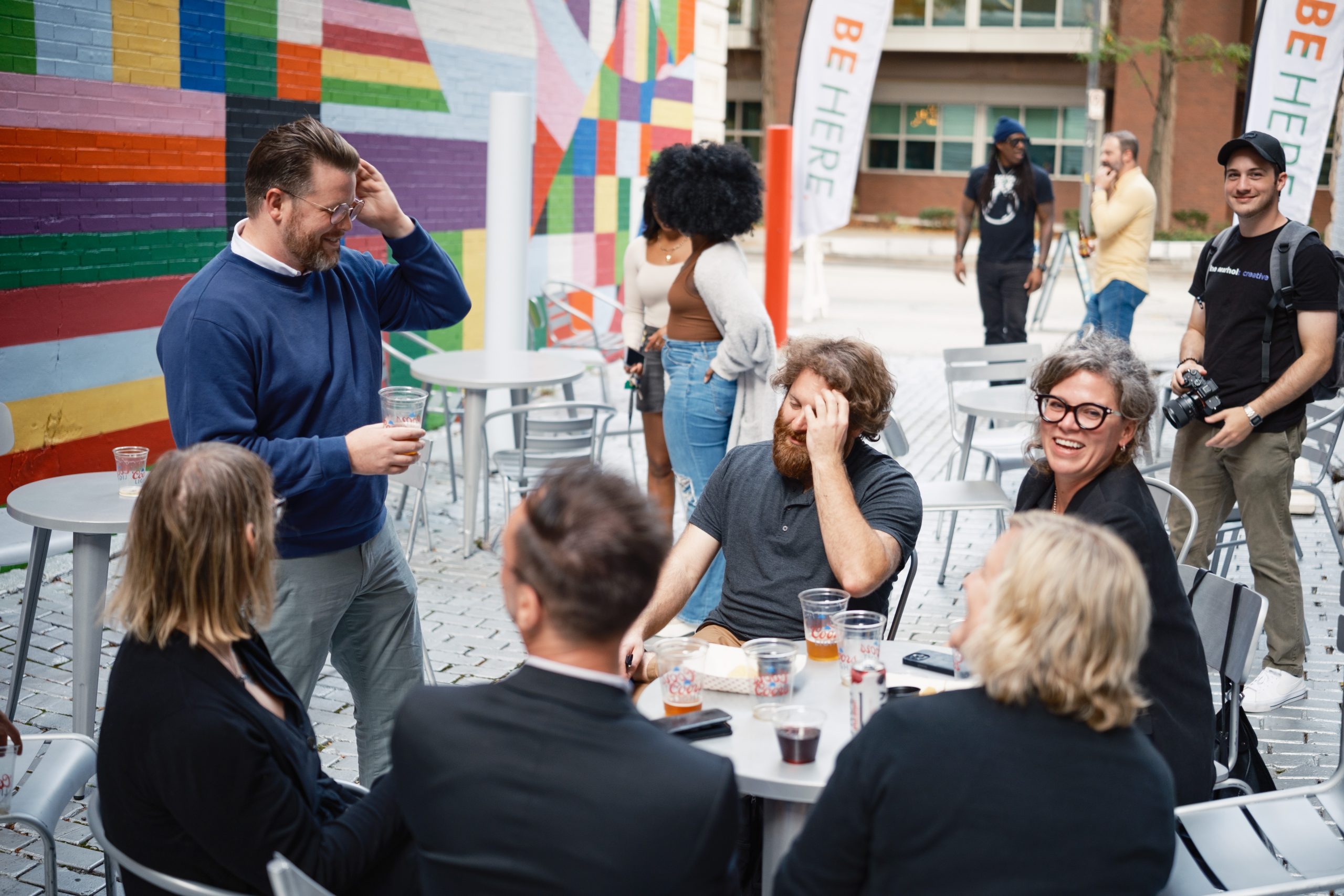 A group of people are gathered around outdoor tables, engaging in conversation and laughter. One man in a blue sweater stands with a drink, scratching his head. Another man with a beard, sitting at the table, also has a hand on his head. A woman with glasses smiles broadly. In the background, there is a colorful mural on a brick wall and a couple of banners that read "BE HERE." Some people are standing, and one person is holding a camera. The atmosphere appears casual and friendly.