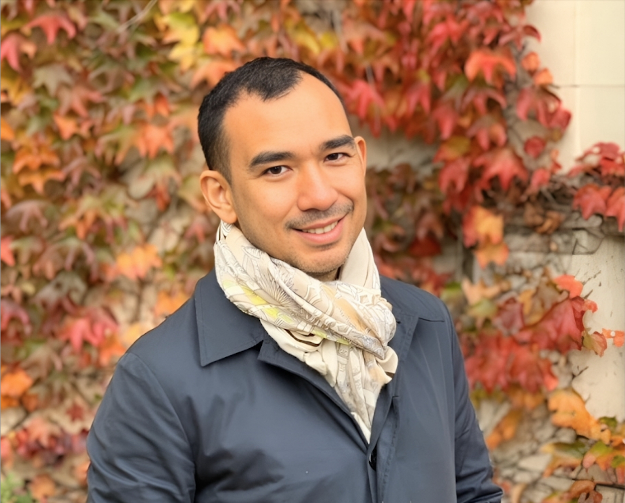 A man in beige scarf and grey jacket standing before a wall covered with red leaves