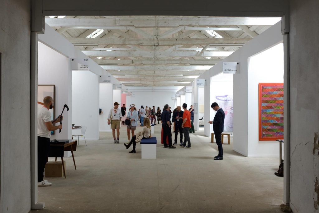 a large hall like space with lots of different booths where art shows are being exhibited, the space is painted white and the people milling around are smartly dressed