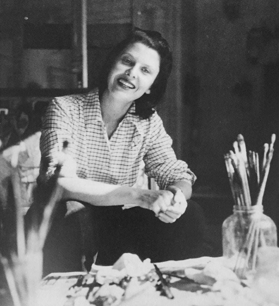 a black and white photograph of a smiling woman sitting before a table covered in paintbrushes and other painting matter