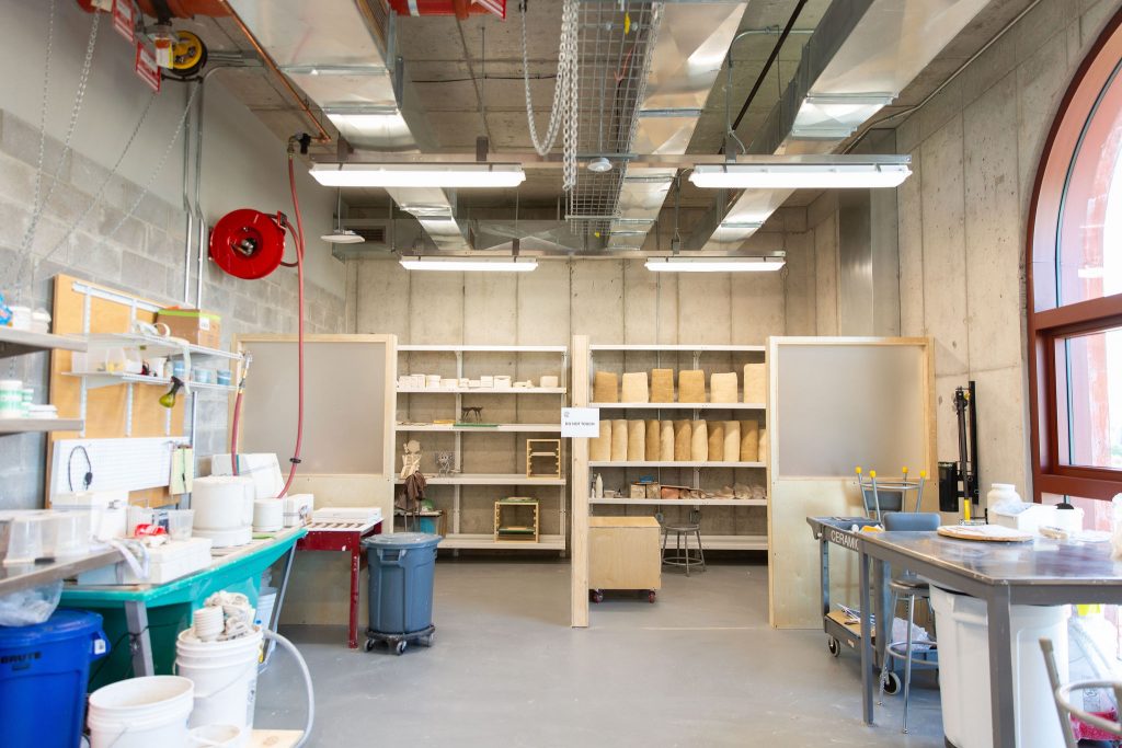 A ceramics fabrication workspace featuring shelves filled with various ceramic pieces and materials. The room is organized with tools, equipment, and supplies neatly arranged. The space is well-lit with large windows and industrial lighting, and it has high ceilings with exposed ductwork and pipes. A sign on one of the shelves reads "DO NOT TOUCH."