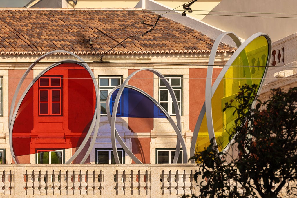 an old orange European building with a large balcony terrace has a sculptural installation installed in which three large oval strutures are filled with glass in red, blue, and yellow that play with the light