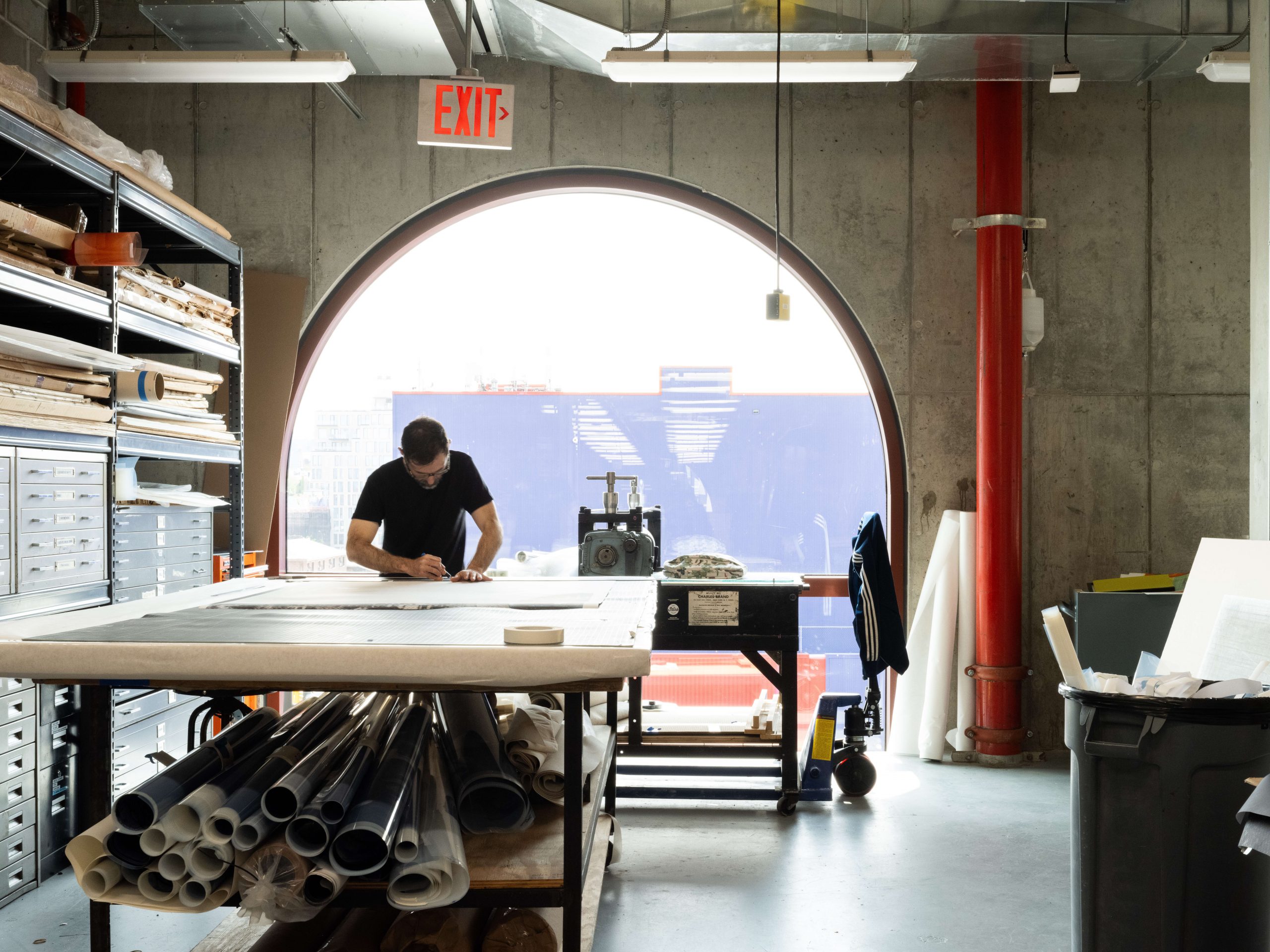 A person is diligently working at a large table in a print shop. The room features a prominent arched window that allows natural light to flood the workspace. The person is surrounded by shelves filled with various tools, materials, and rolled prints. The industrial aesthetic is highlighted by concrete walls, exposed pipes, and a red pillar. The environment is organized and well-equipped for detailed printmaking work.