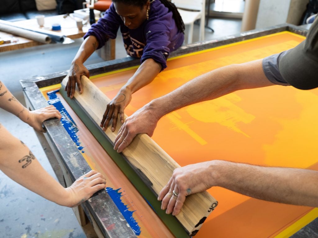A group of people are collaboratively working on a screen printing project. They are using a large squeegee to spread ink over a screen with a bright orange background. The workspace is equipped with tools and materials necessary for screen printing. The scene captures the hands-on and cooperative nature of the activity, with several hands visible in the process of creating the print.