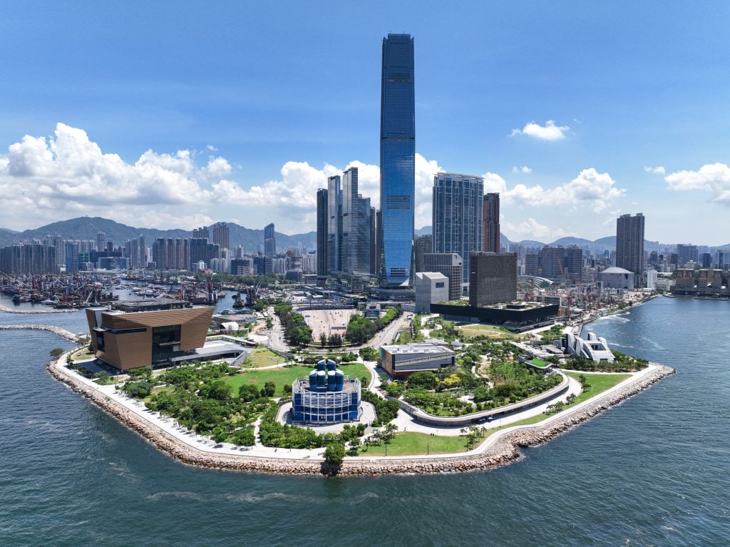 an aerial photo of west kowloon in hong kong. the image features tall buildings surrounded by water and blue skies