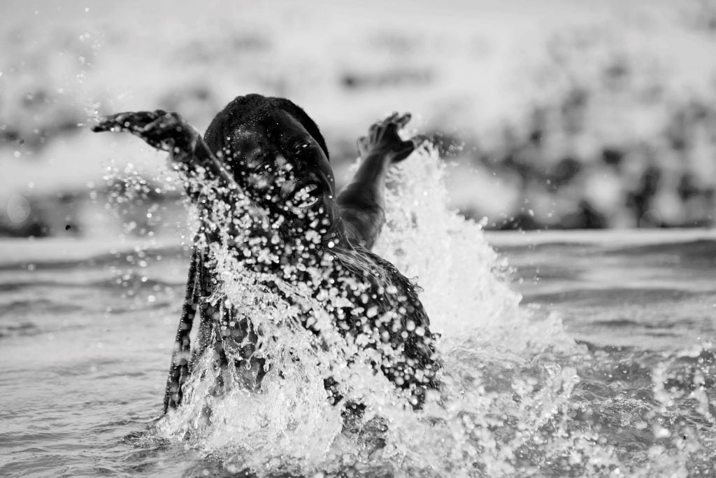 a black and white photo of a person splashing in water, arms outstretched. By artist Zanele Muholi.
