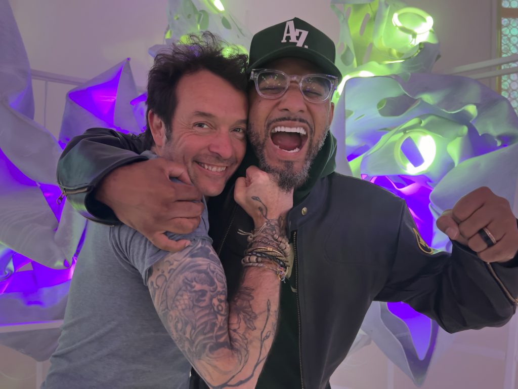 The artist Arne Quinze and musician Swizz Beatz hug in front of a white sculpture with wide smiles.
