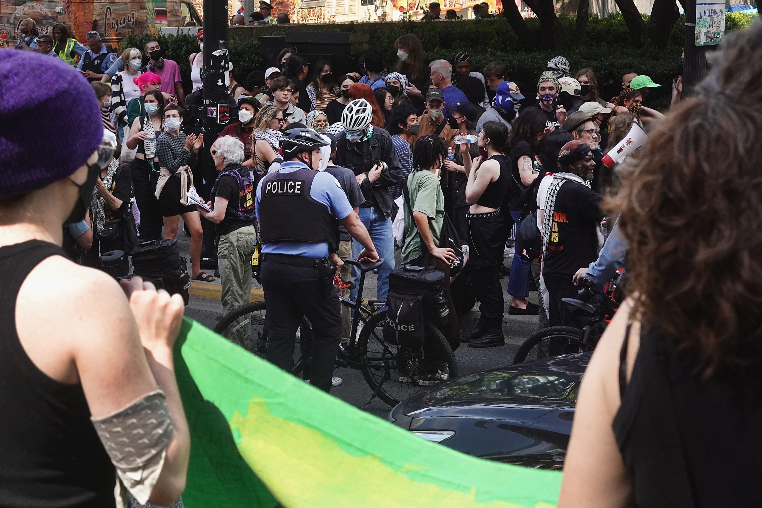 A horde of people gathering outside a museum, with police officers on the perimeter of the crowd. Two people hold a Palestinian flag in the foreground