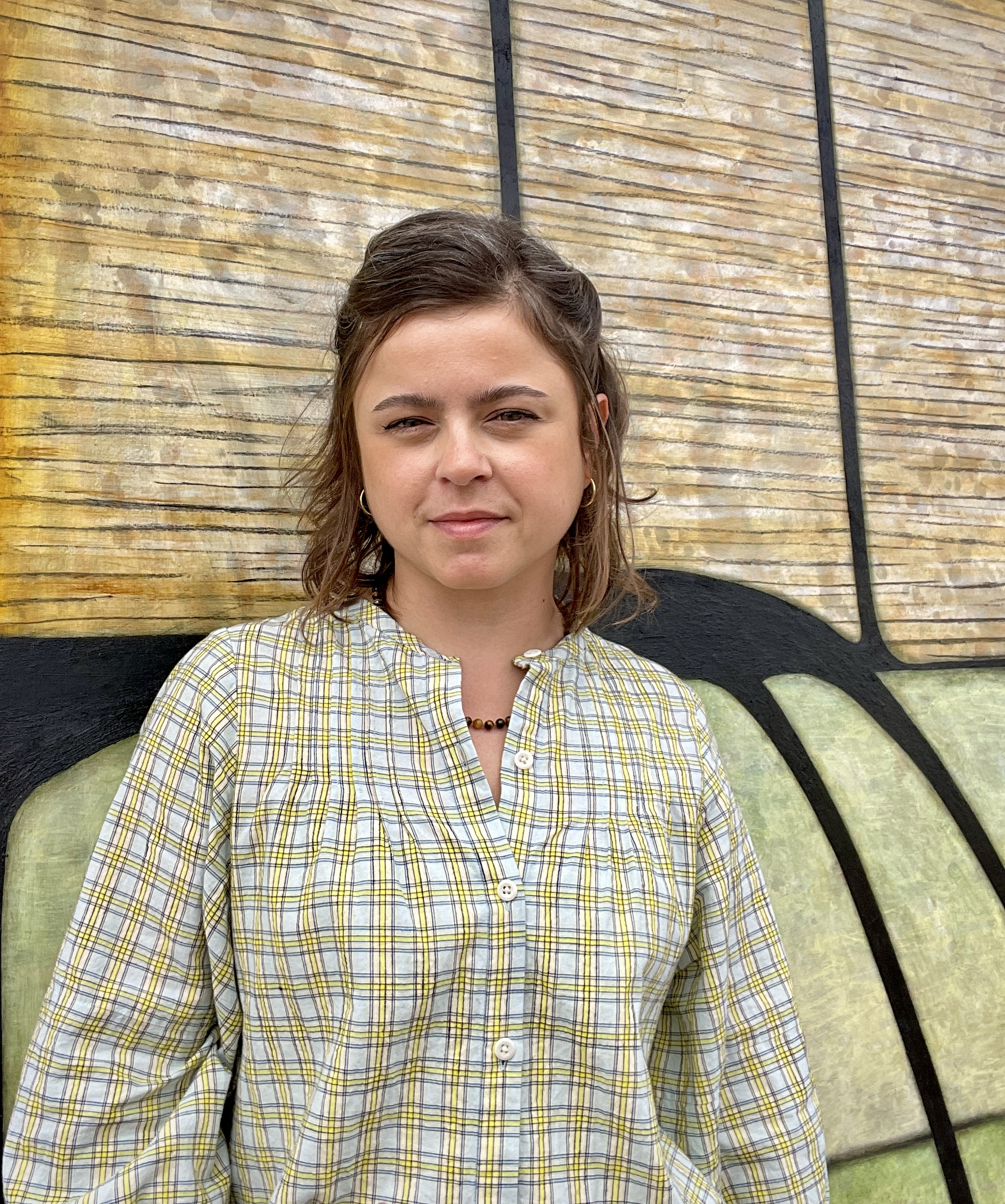 Portrait photo of the artist Gabriela Giroletti wearing a beige and white plaid peasant shirt standing in front of one of her abstract canvases in earthy tones.