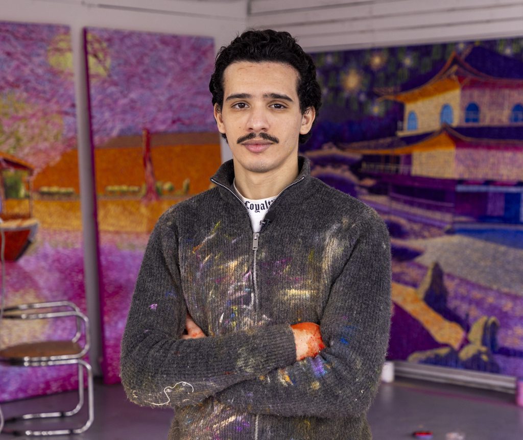 Portrait of artist Touils standing in front of a selection of his canvases in the studio with his arms crossed, and zip up hoodie covered in paint marks.
