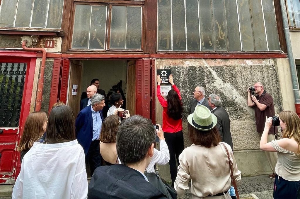 A group of people, including photographers and onlookers, are gathered outside an old building with large windows. A woman in a red top is affixing a plaque to the wall next to the entrance, with several people observing and taking pictures. The building appears to be in a state of disrepair, with weathered and stained walls. The number "11" is visible above the entrance.