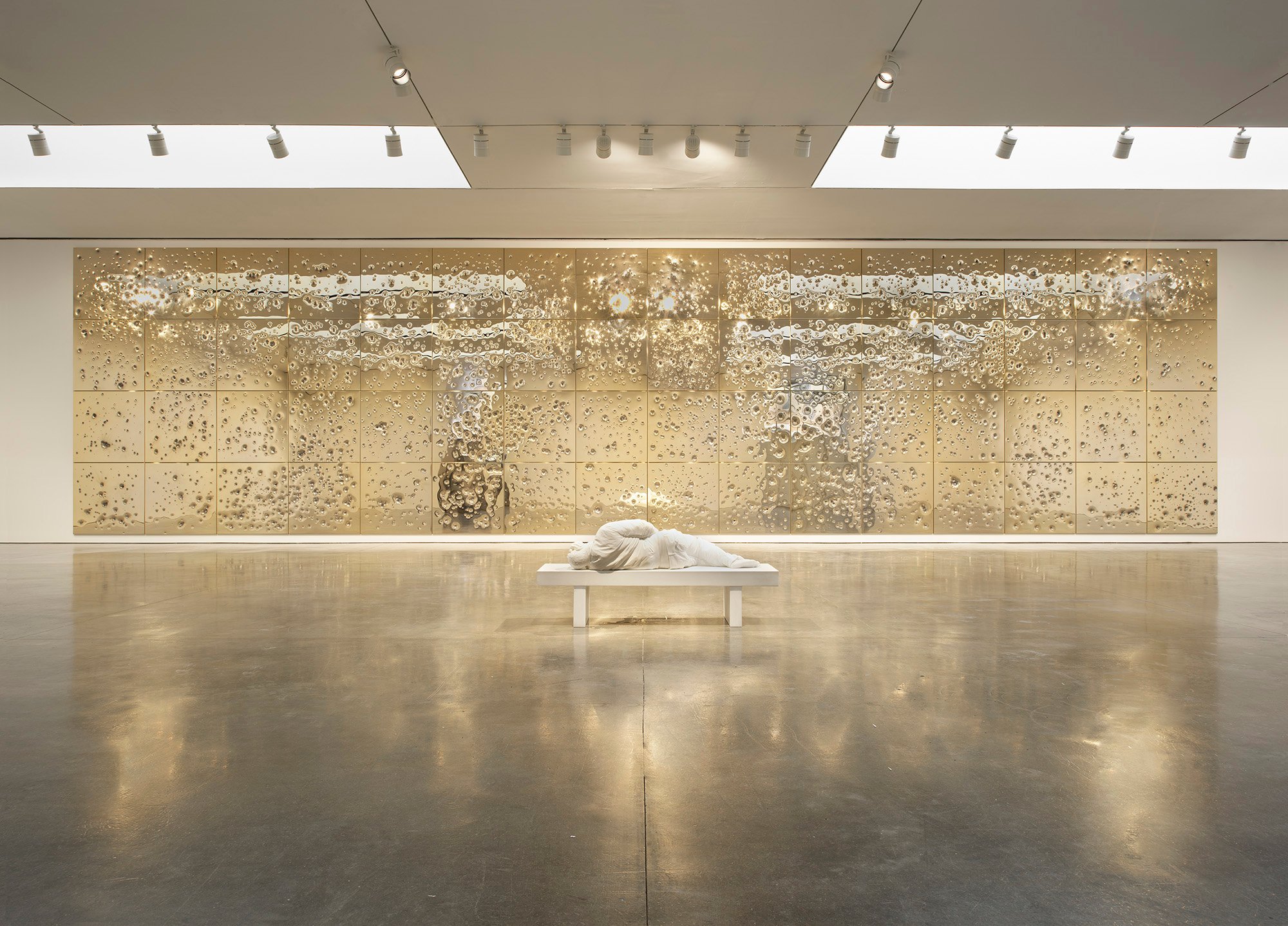 A sculpture of a man lying on a bench is positioned in the middle of a galley, against a wall of metallic panels pockmarked with bullet holes.