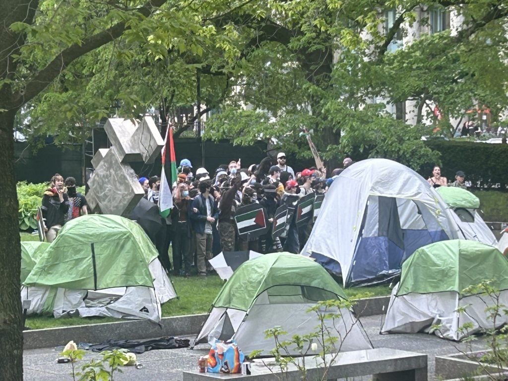 A scene of a protest in an urban park, with demonstrators gathered among tents. Some individuals are holding flags, and the group appears engaged and vocal. The setting includes trees and a large abstract sculpture, contributing to a dynamic and populated atmosphere.