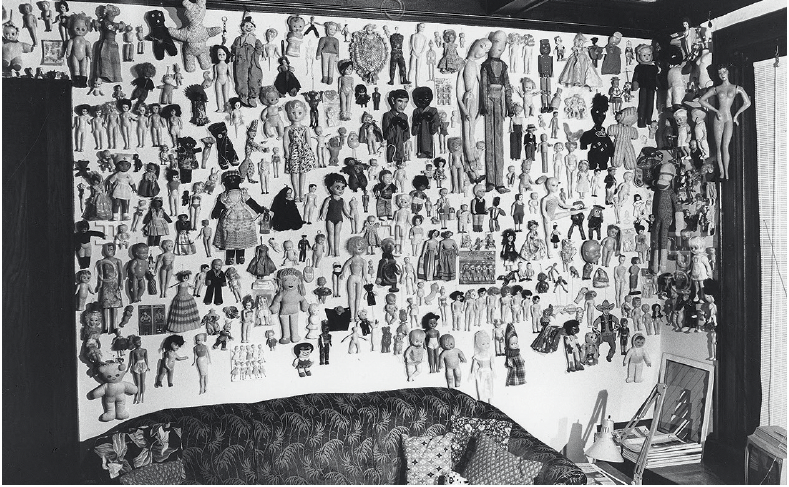 A blac-and-white photo of inside Christina Ramberg's Chicago Apartment from 1972 showing hundreds of different types of doll mounted on the wall above a tufted sofa.