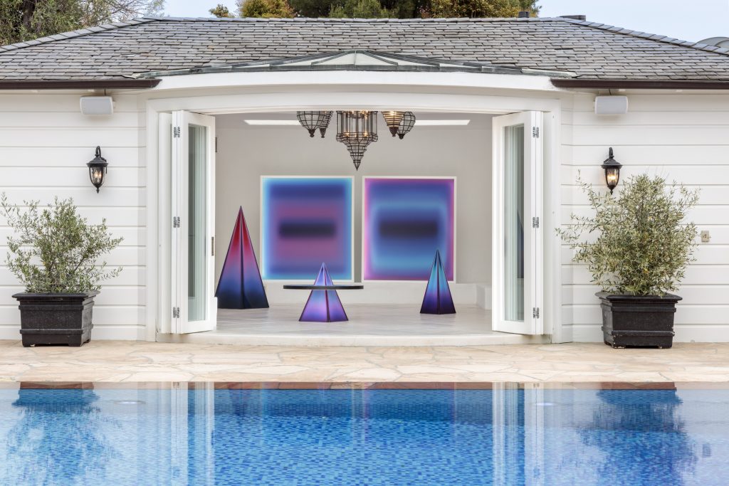 Some ombre purple pyramid sculptures and two paintings hang in a colonial mansion in front of an electric blue swimming pool
