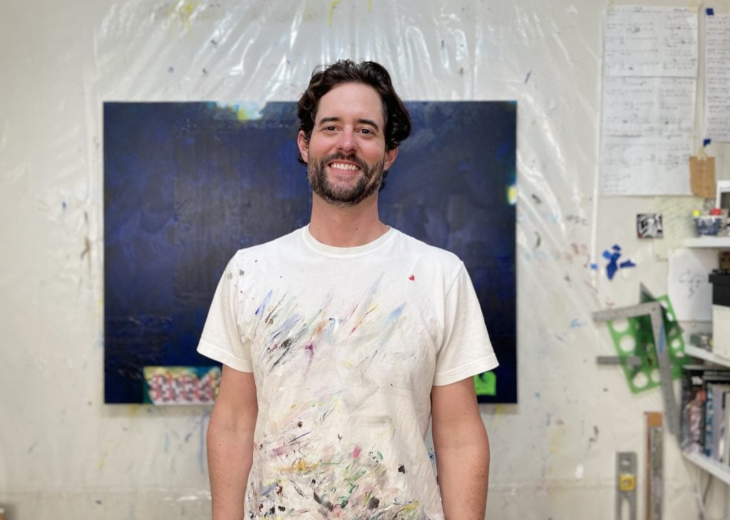 a man in a white paint splattered tee shirt stands in front of a blue painting