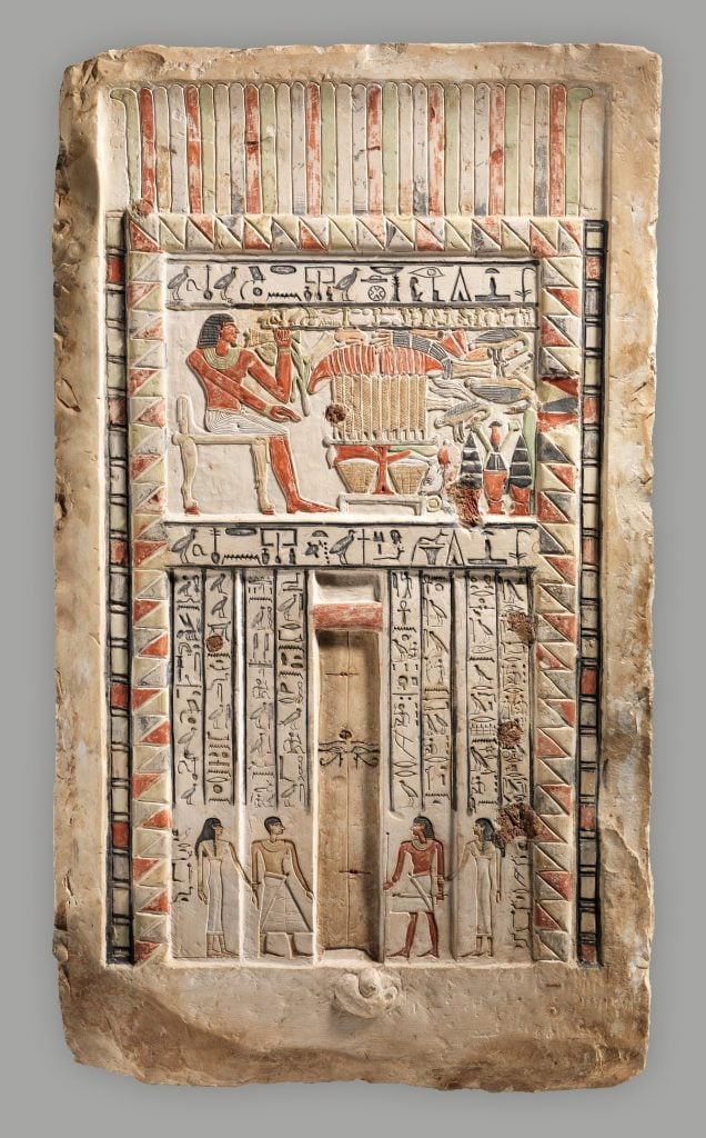 An ancient Egyptian limestone stela with a large carving of a man seated at a table loaded with a feast, and inscriptions beneath him.